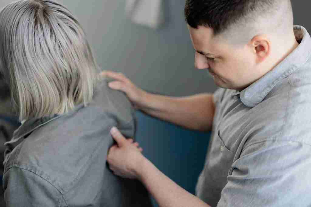 A man adjusts the back of a woman's jacket ensuring a proper fit as she stands in a casual setting